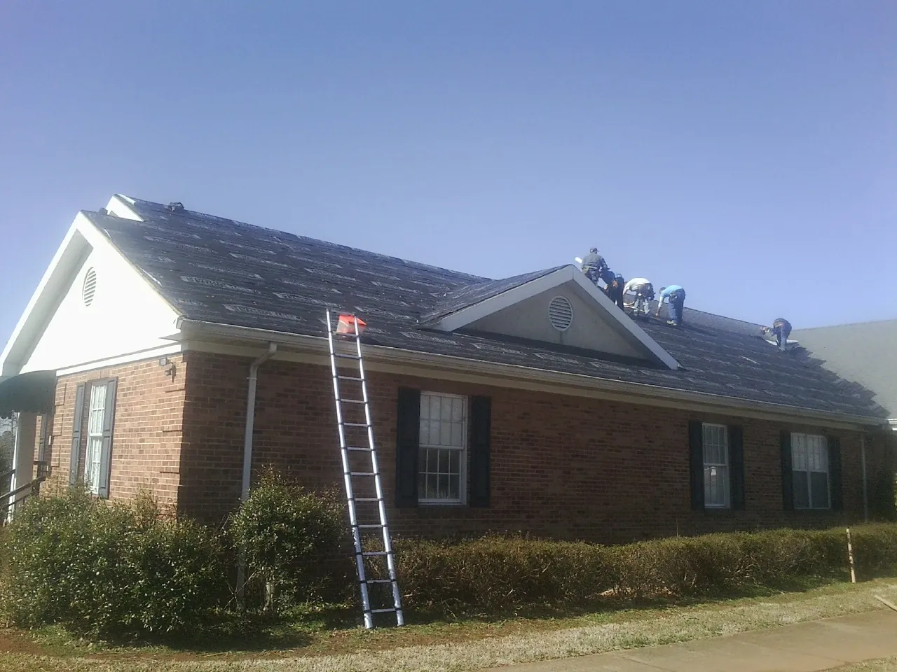 Crew tearing shingles off a brick ranch roof with a ladder set — Emergency Roof Repair work by our crew in Saint Regis