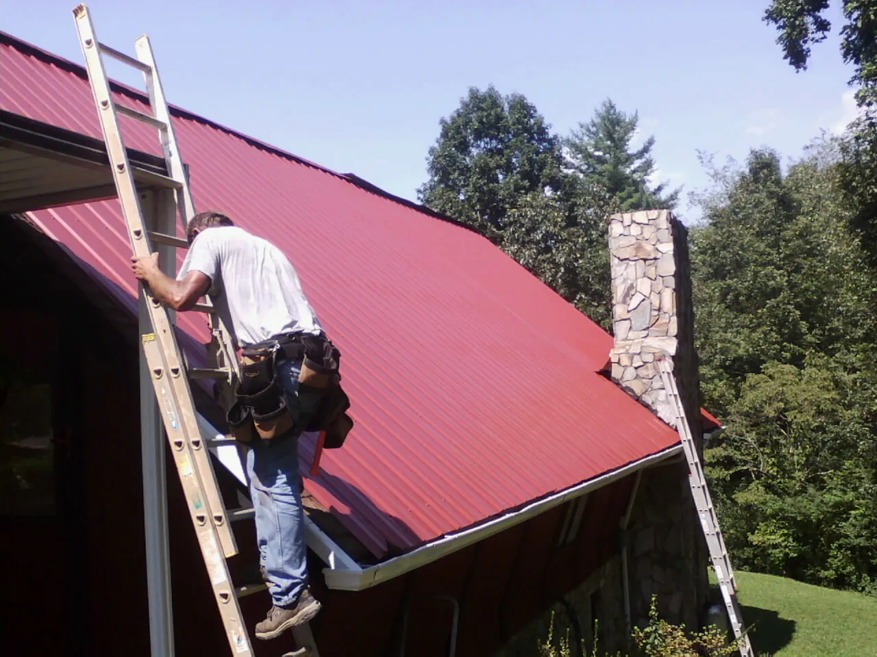 Roofer on a ladder beside a red metal roof and stone chimney — Soffit & Fascia Repair by Roofing Brothers in Saint Regis